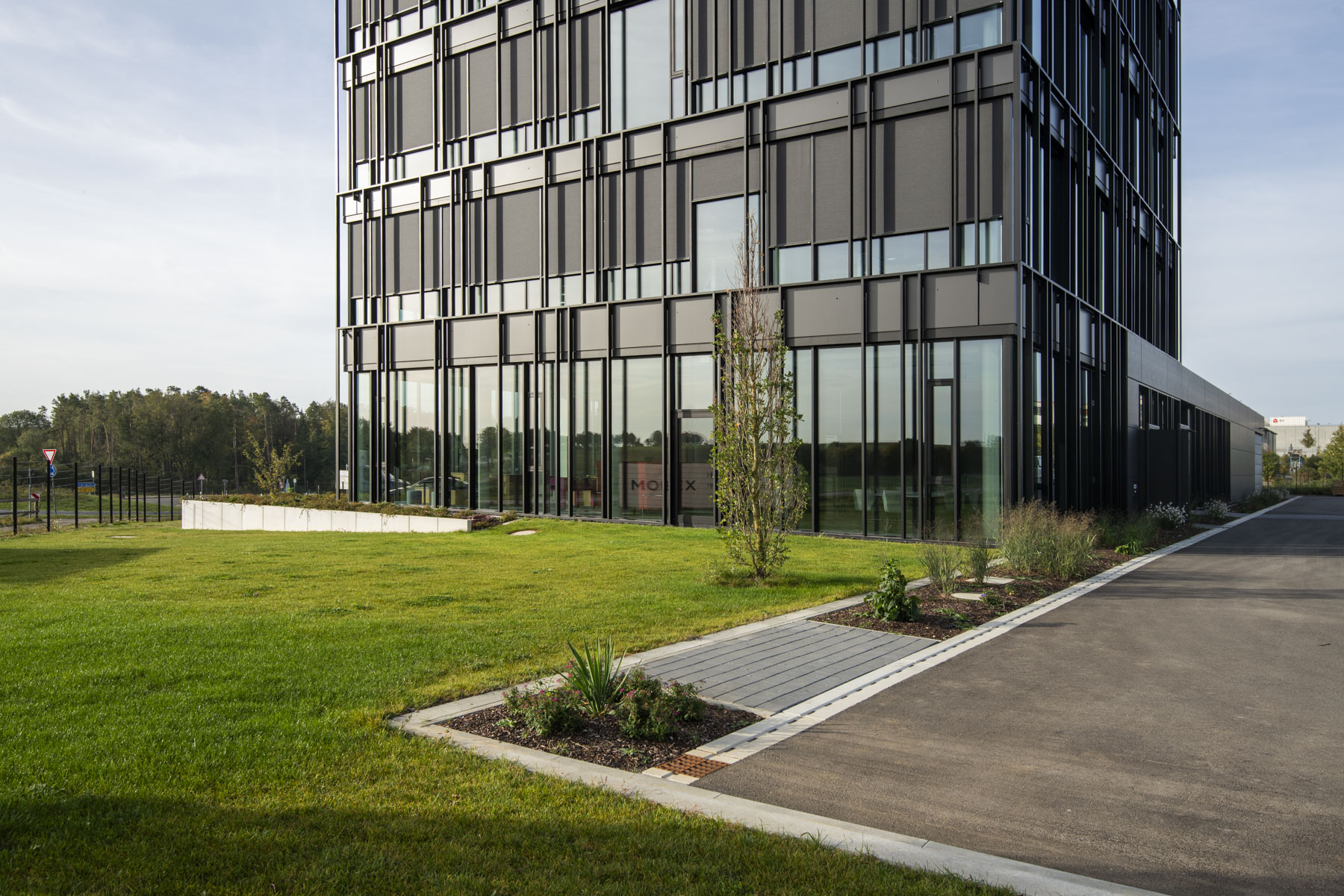 Mehrstöckiges schwarzes Firmengebäude mit großzügigen Glasflächen, davor grüner Rasen, frisch bepflanzte Grünflächen und ein asphaltierter Parkplatz