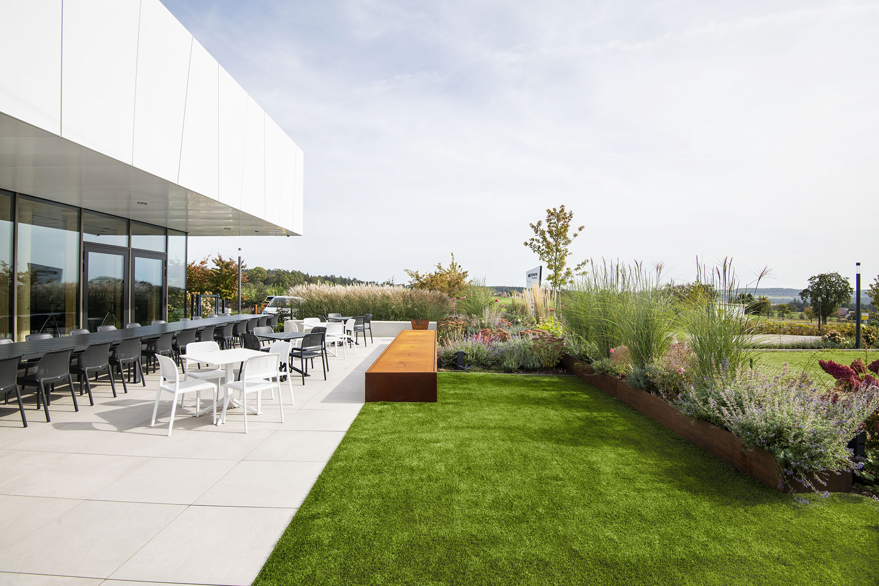Bestuhlte Terrasse mit grüner Wiese, braunem Wasserspiel und hochstehenden Blühpflanzen